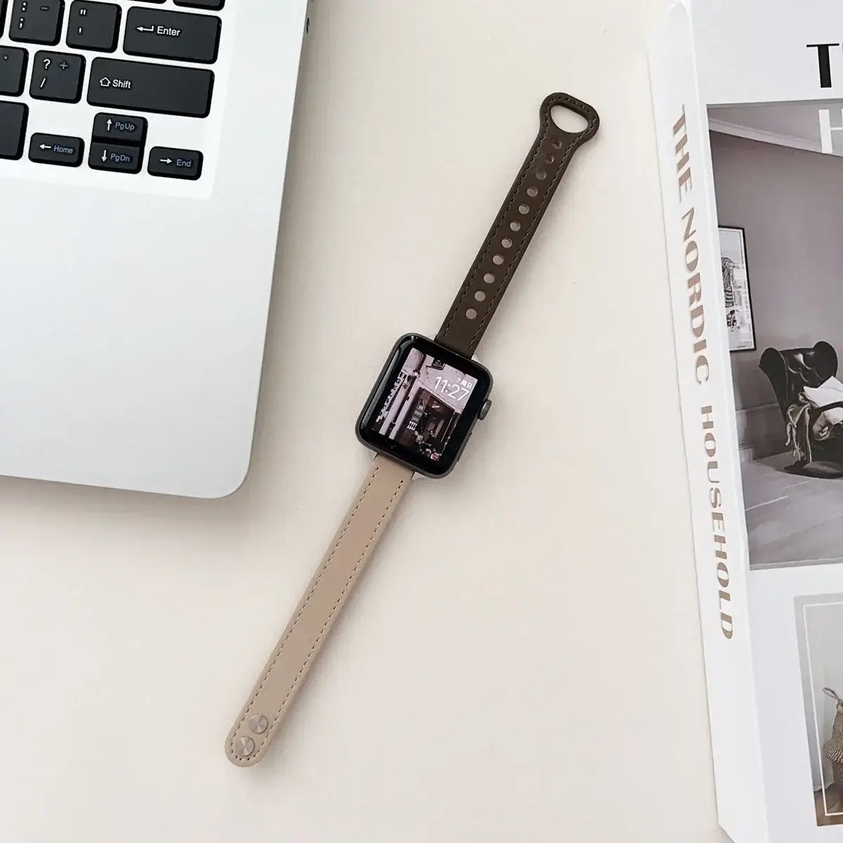 Thin leather Apple Watch band in beige and brown, resting next to a laptop and magazine on a minimalist desk.
