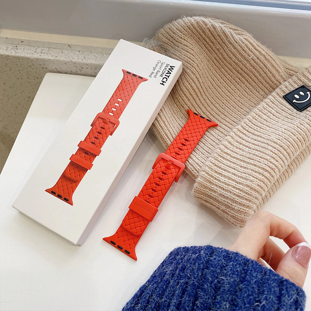 Apple Watch silicone strap in red color with braided design, next to a beige knit hat and packaging.