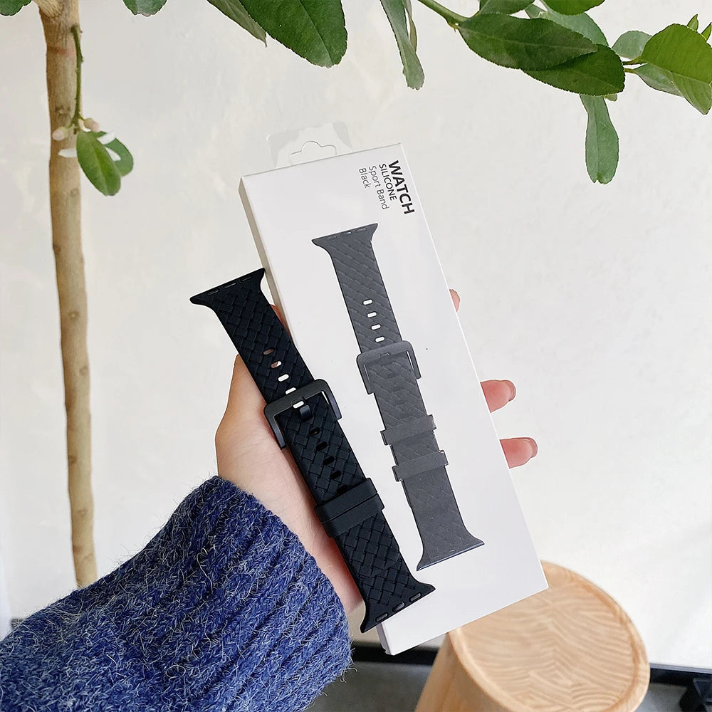 Black silicone Apple Watch strap showcased in packaging, held in hand against a home interior backdrop.